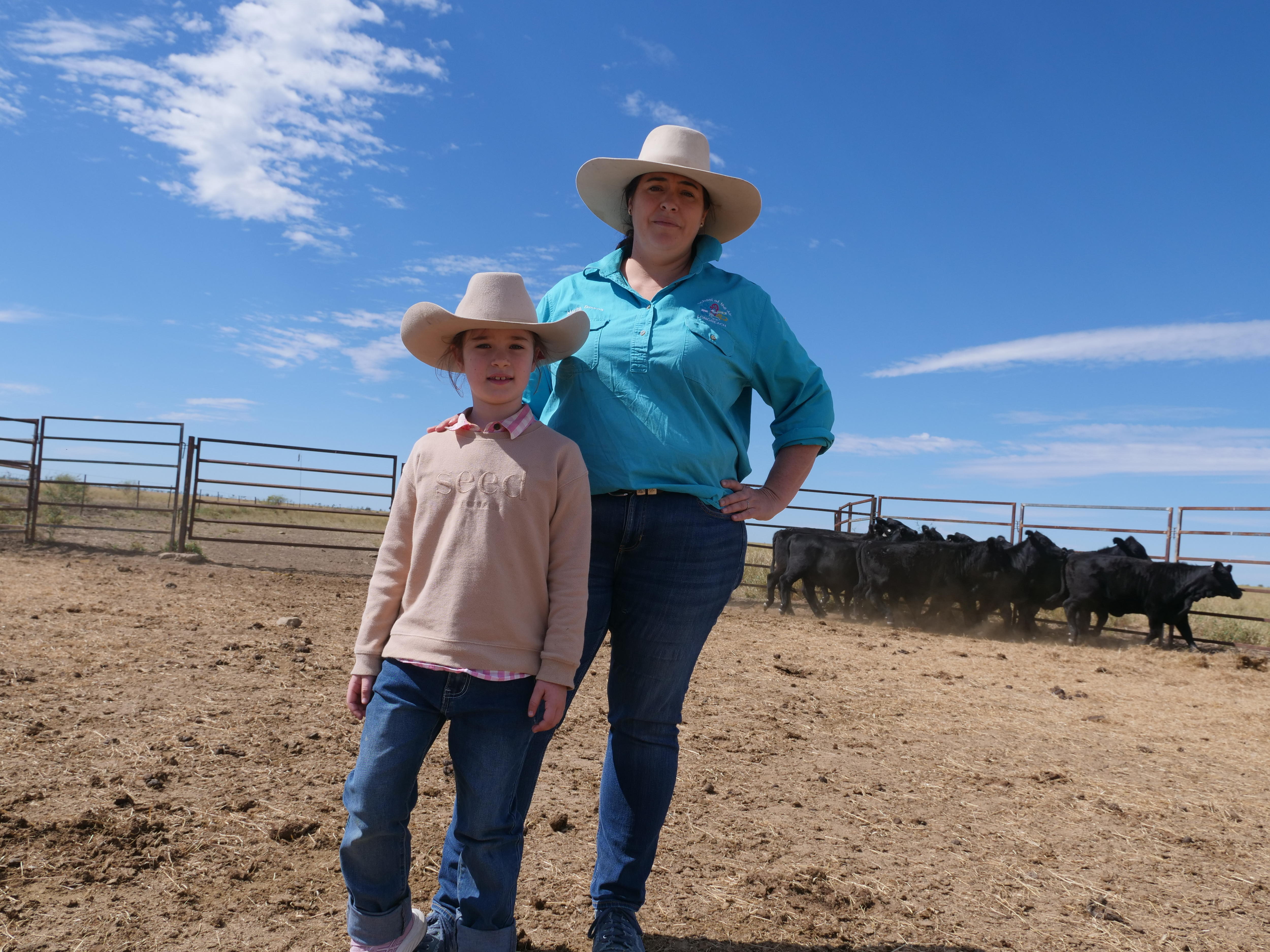 A mother and her daughter in akuburas stand with confidence in a stockyard with a mob of cattle running behind them.