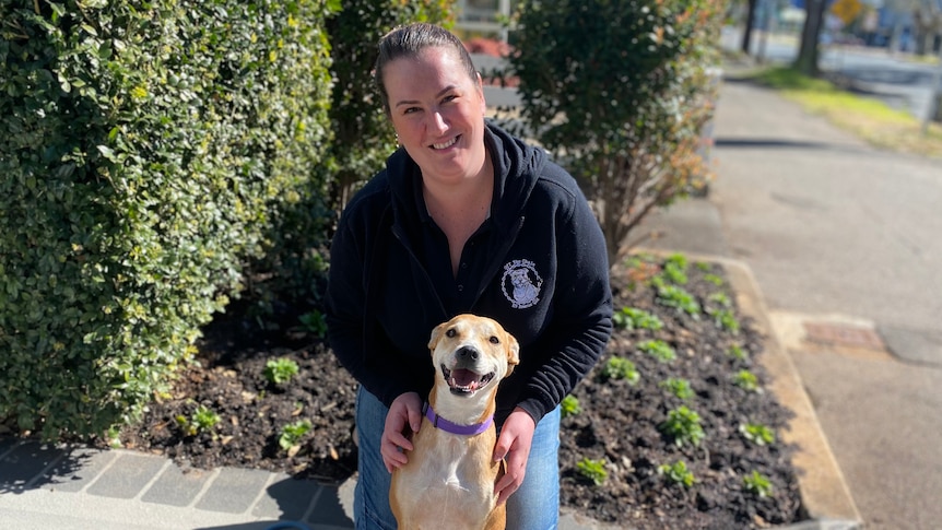 A smiling woman crouches behind a pale-coloured dog that sits in front of her.