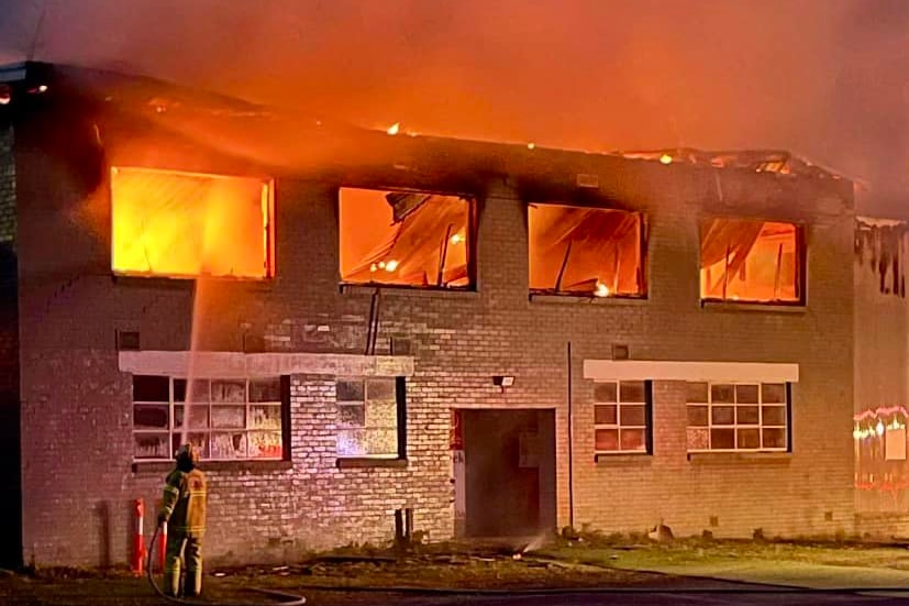 A roof has collapsed and flames can be seen inside a two storey brick building as a lone firefighter sprays water into a window