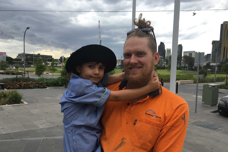 A man in a bright orange top, with his hair in a bun, holds his daughter who wears a school uniform and hat.
