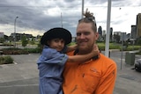A man in a bright orange top, with his hair in a bun, holds his daughter who wears a school uniform and hat.