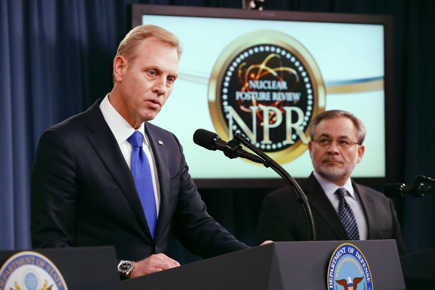 A US defence official looks on as another speaks into a microphone at a podium.