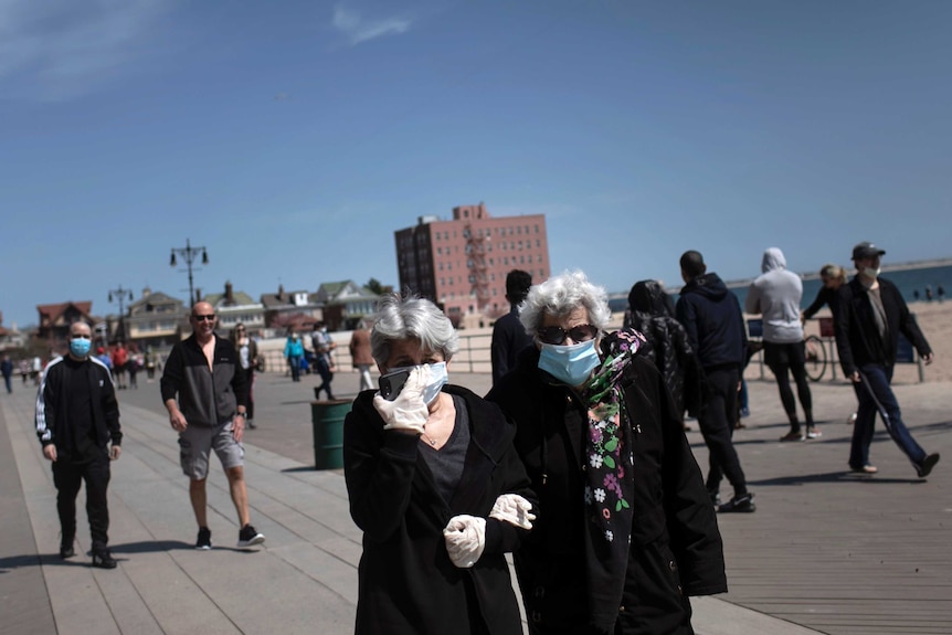 Two older women wear masks as they walk on a beach boardwalk.