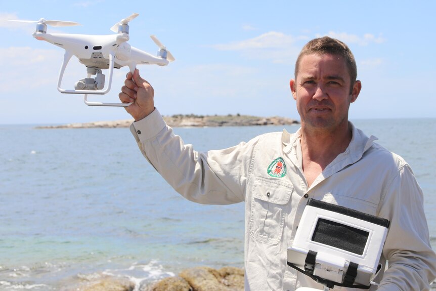 Man holding display unit and holding up white drone, on coast with island in background