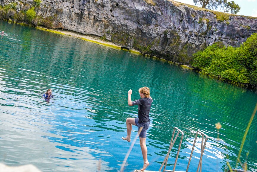 A boy wearing bathers jumps off a pontoon into a large lake of water surrounded by limestone walls.