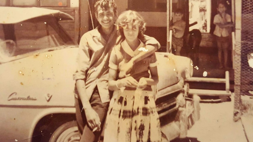 An old sepia-toned black and white photo of a young man and woman posing in front of a car.