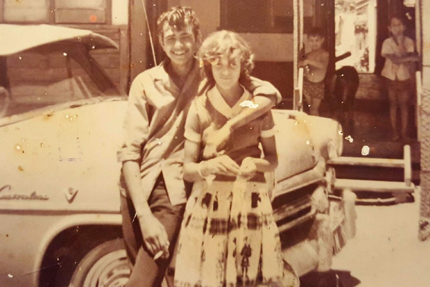 An old sepia-toned black and white photo of a young man and woman posing in front of a car.