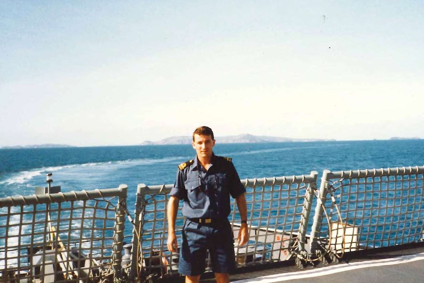 Man standing on the side of ship in a blue navy uniform with ocean in background