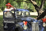 A woman wearing a red hat hangs outside the open door of a vintage car and waves