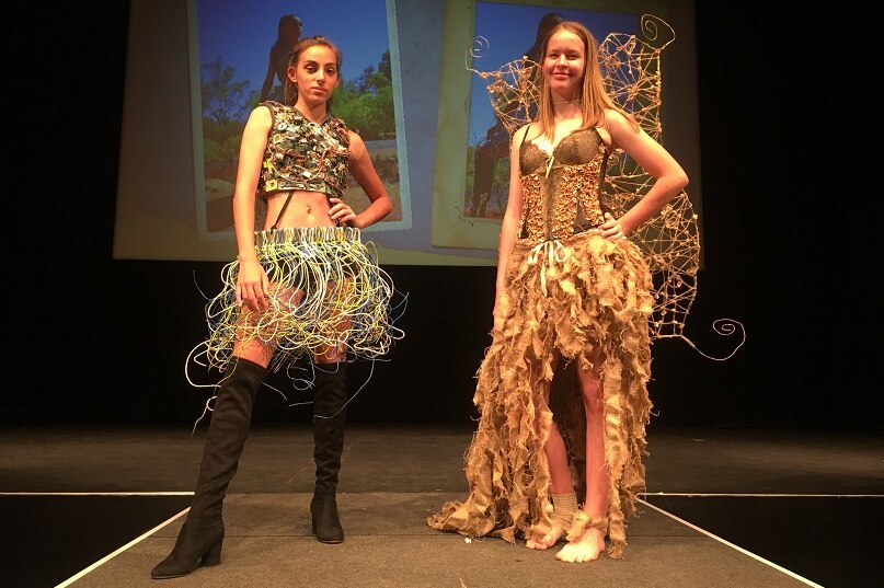 Two models stand on a catwalk wearing artistic designs