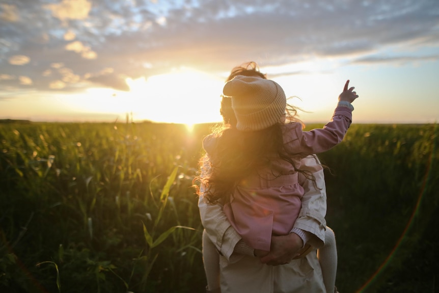 Adult holds young child on back while walking through a green field at sunset. 