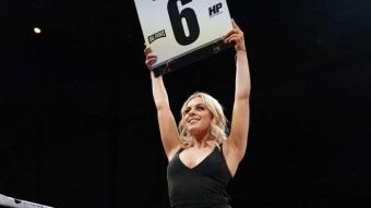 A blonde woman in a black evening dress holds a round card in a boxing ring