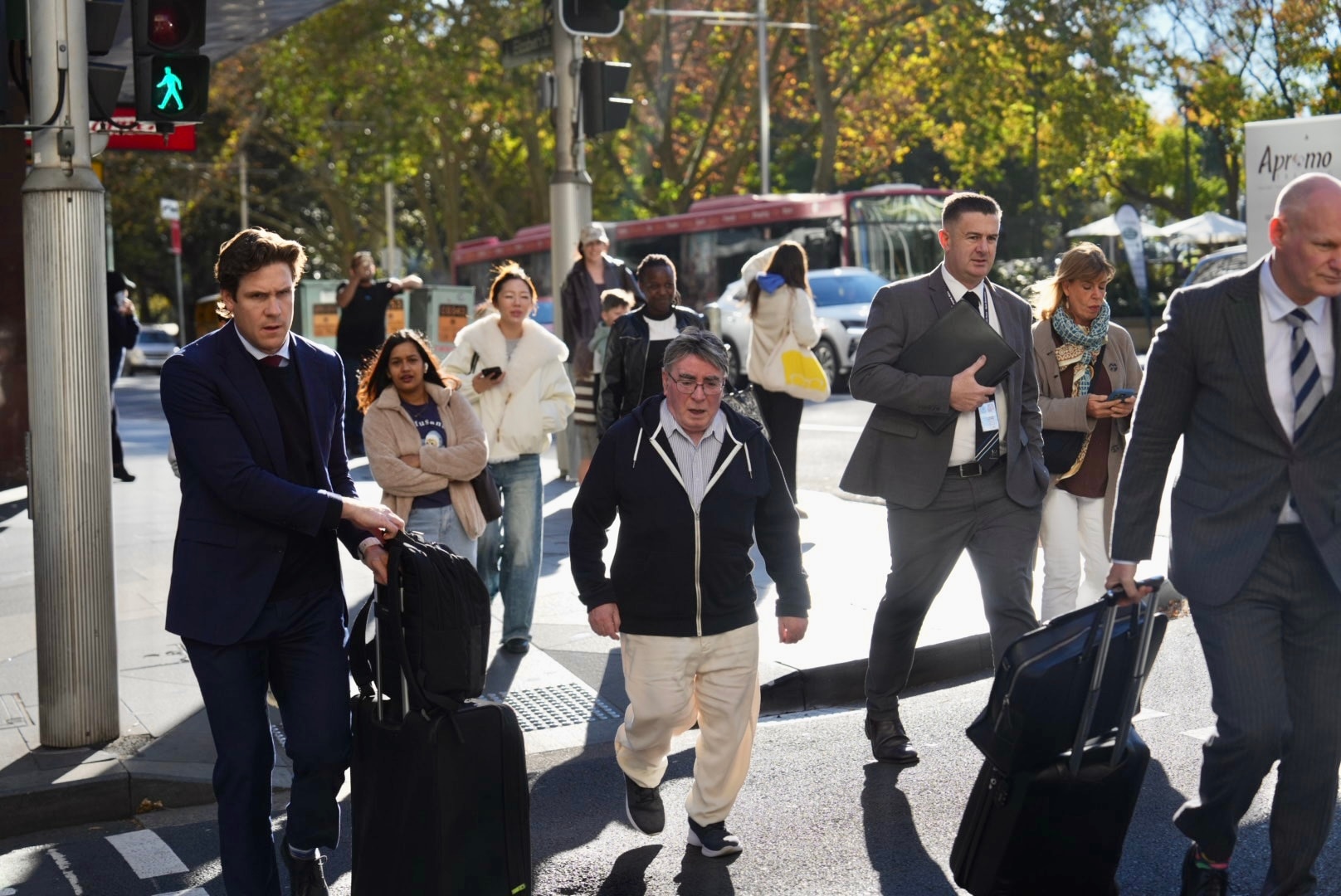 small grey-haired man in chinos and jumper crossing street surrounded by men in suits with suitcases 