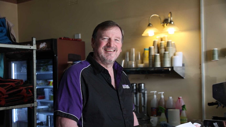 Middle aged man wearing a black and purple polo shirt standing at a cash register, low light photo.