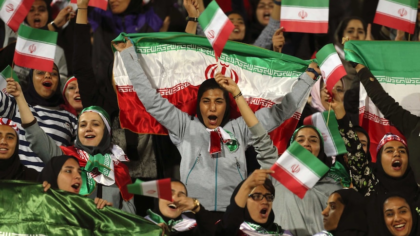 Women wearing headscarfs hold Iranian flags and cheer.