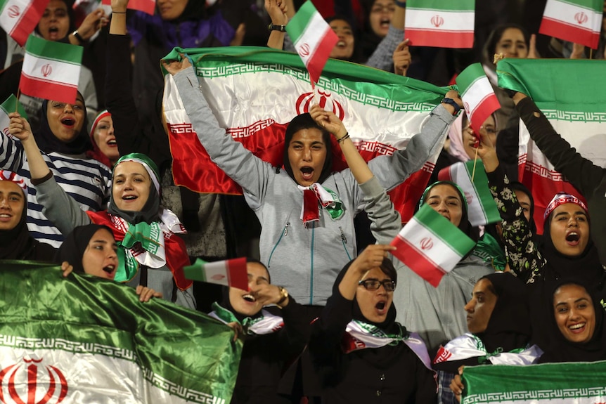Women wearing headscarfs hold Iranian flags and cheer.