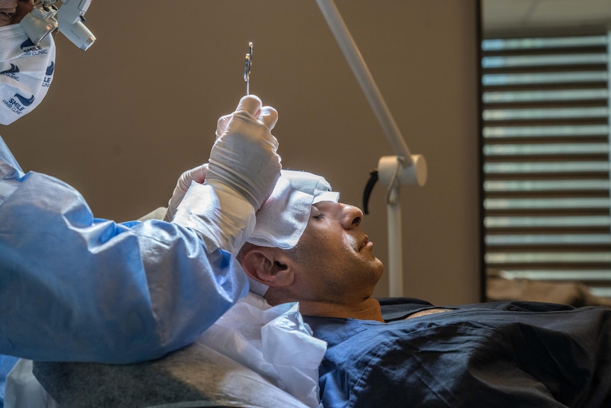 A man wearing blue hospital gown lays back while a doctor in scrubs uses an instrument on the patient's scalp