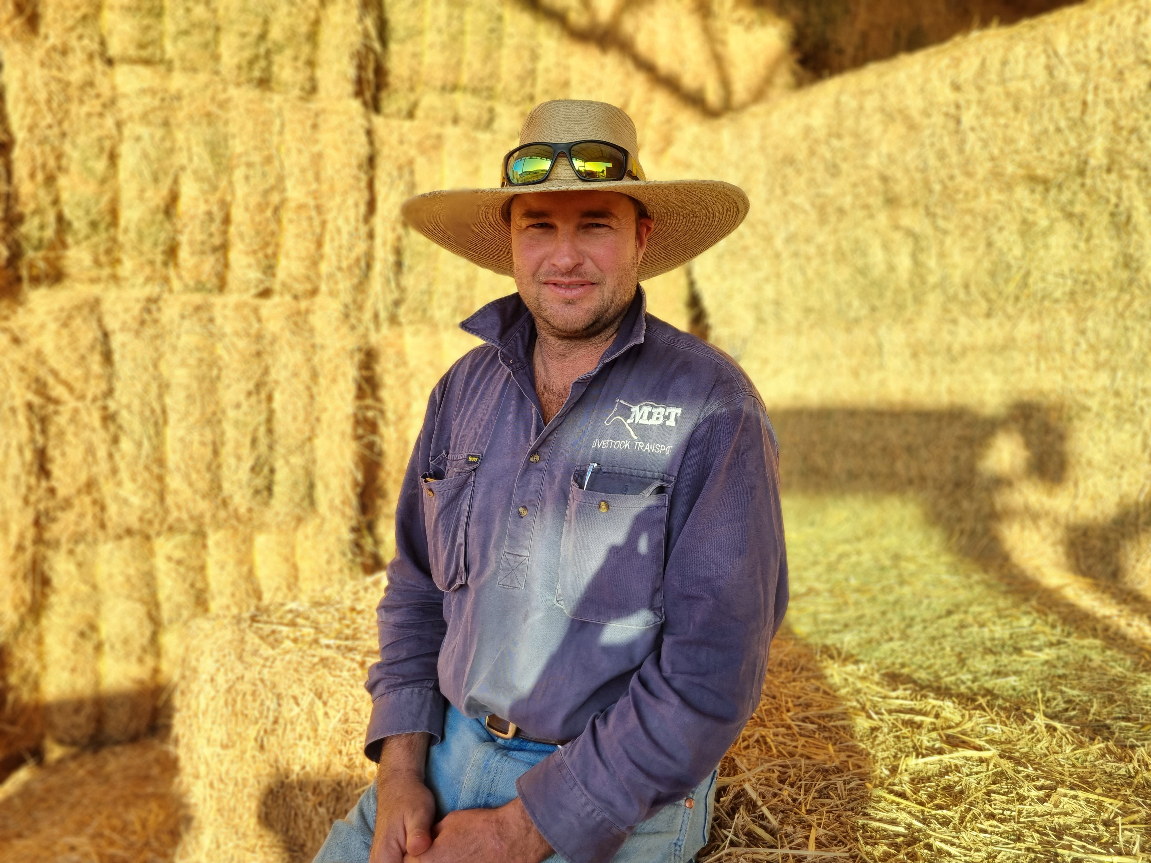 A man in a hat and blue shirts sits in a hay shed.