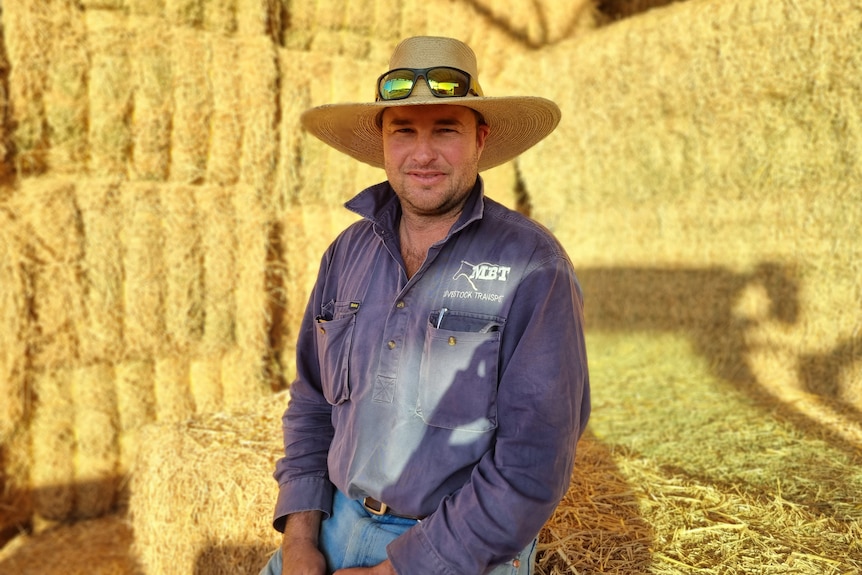 A man in a hat and blue shirts sits in a hay shed.