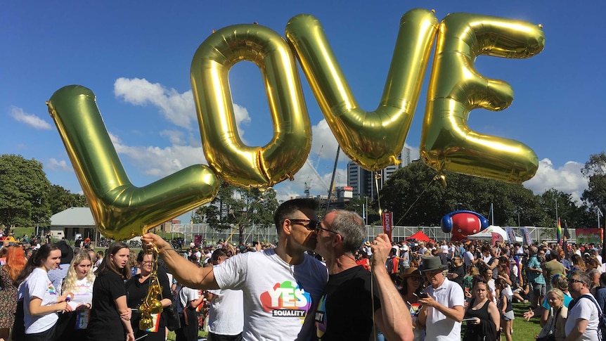 Two men kiss under gold ballons spelling out "LOVE"