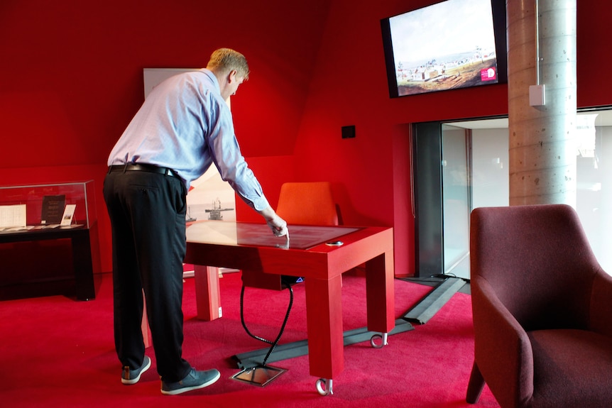 A man stands in front of a red table with an inbuilt interactive screen looking at an historical painting.