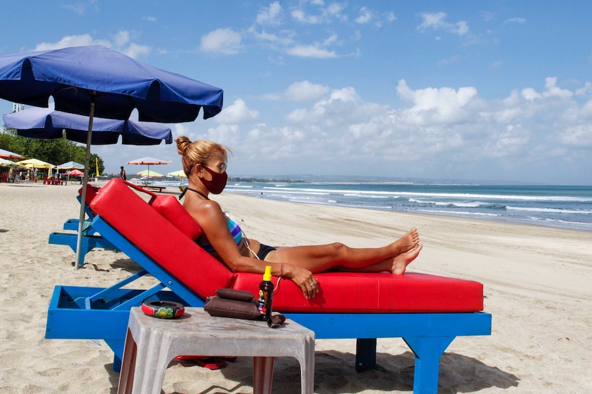 A woman in a bikini and facemask sits on a deck chair on a beach