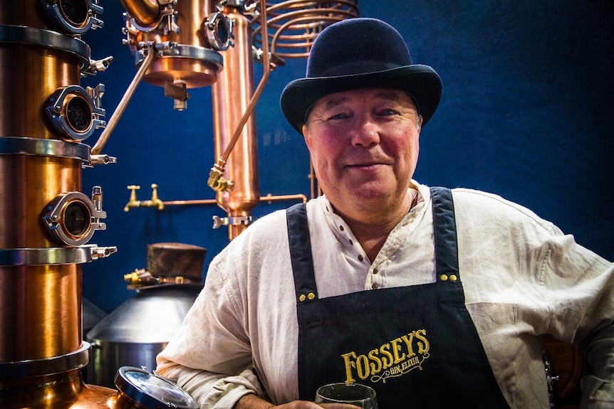 Man in hat and apron in front of copper distillery