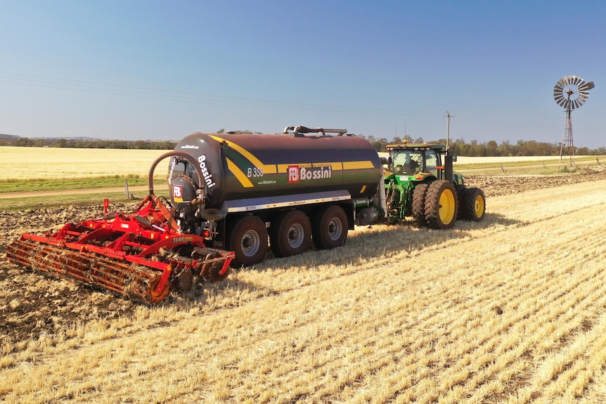 A slurry tanker is driving through a paddock with a windmill in the background.