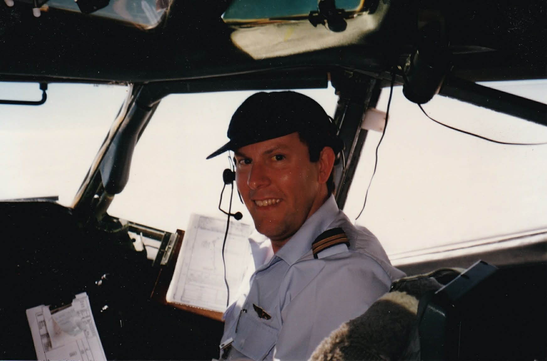 Mr Klouth in a white uniform wearing a cap and headset while sitting in a cockpit smiling.