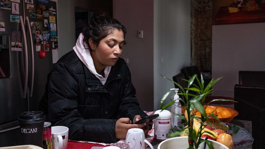 Shayla Shakshi sitting at a kitchen bench looking at her mobile phone