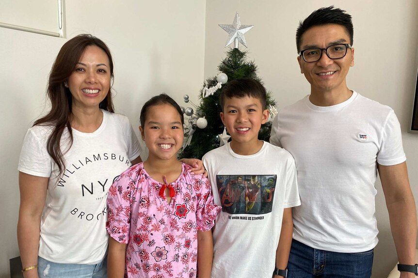 A family of four stand in front of their Christmas tree smiling.