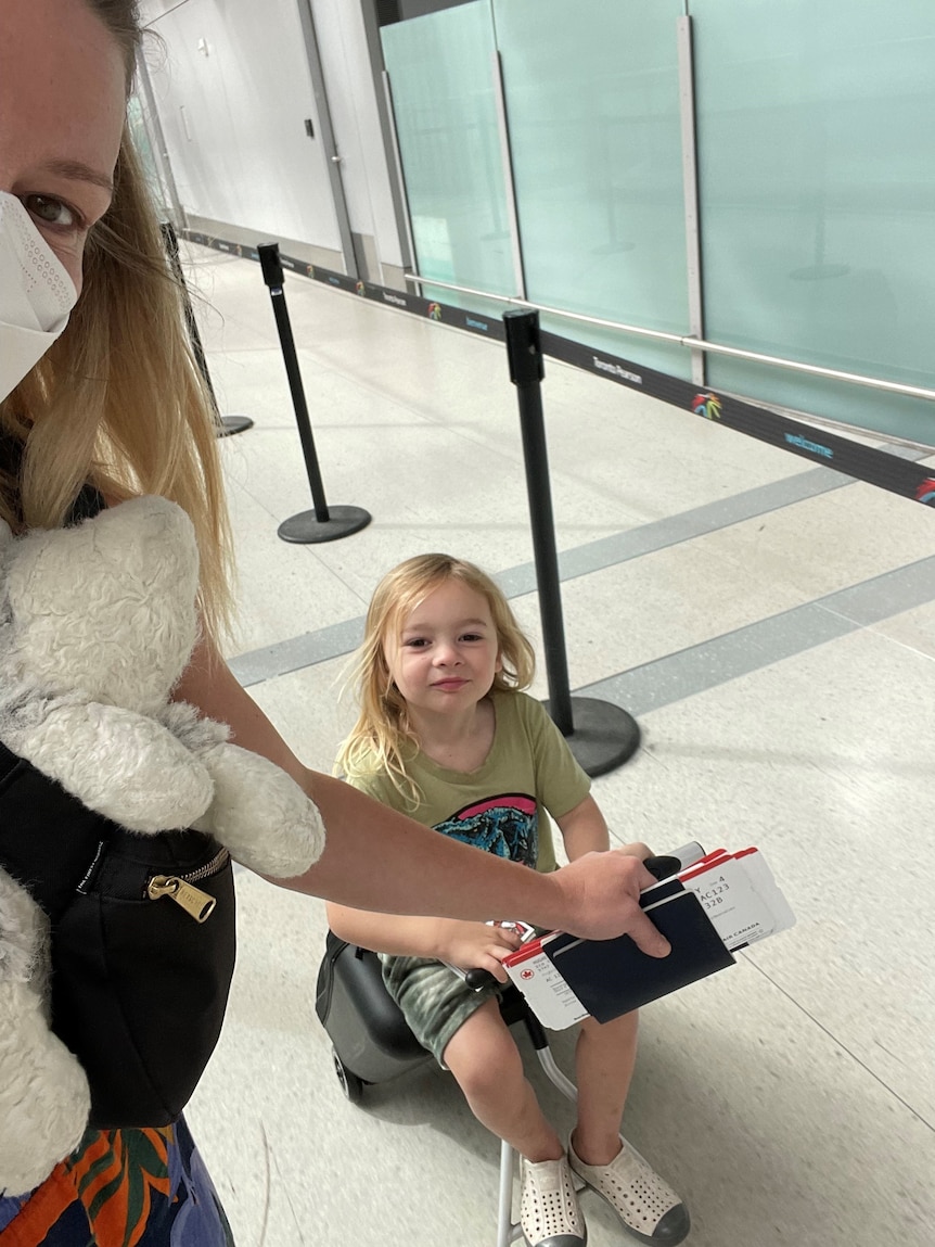 A woman wearing a face mask pulls a young child sitting on a rolling suitcase through an airport.