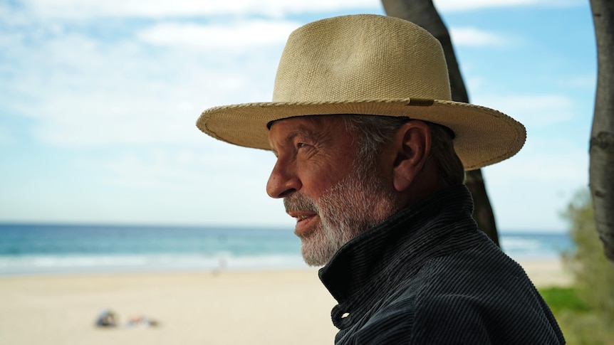 Sam Neill wears a tan wide brim hat, looking out to sea at the beach in profile view