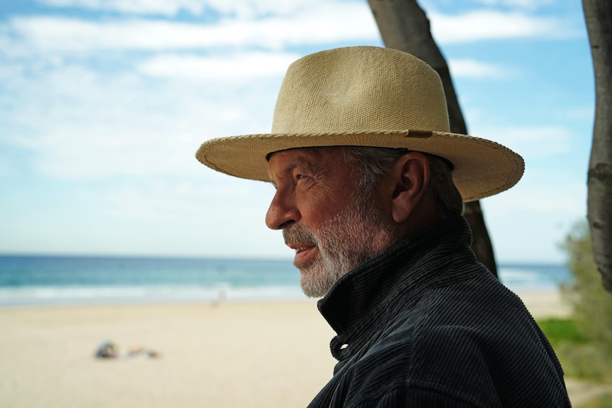 Sam Neill wears a tan wide brim hat, looking out to sea at the beach in profile view
