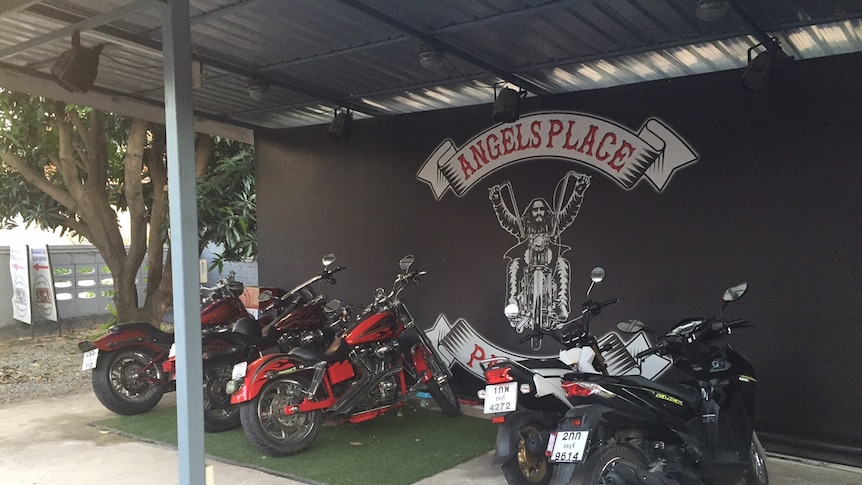 Black wall with 'Angels Place' logo and mural of bikie, with several motorcycles parked in front