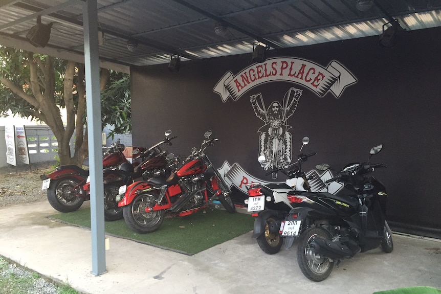 Black wall with 'Angels Place' logo and mural of bikie, with several motorcycles parked in front