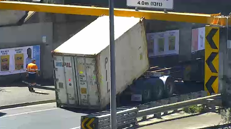 A truck's shipping container load can be seen stuck under a yellow metal beam on a road bridge.
