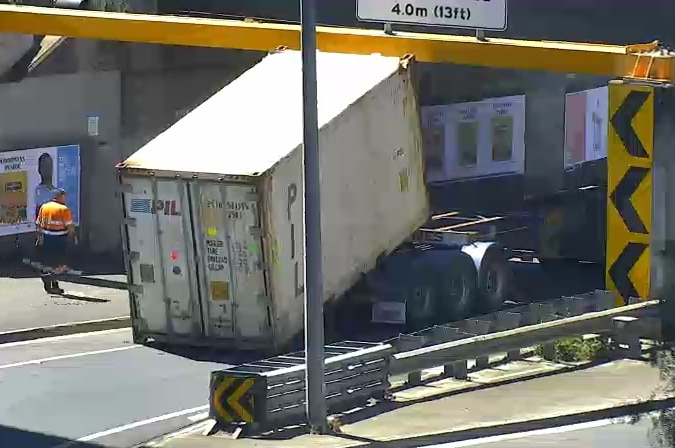 A truck's shipping container load can be seen stuck under a yellow metal beam on a road bridge.
