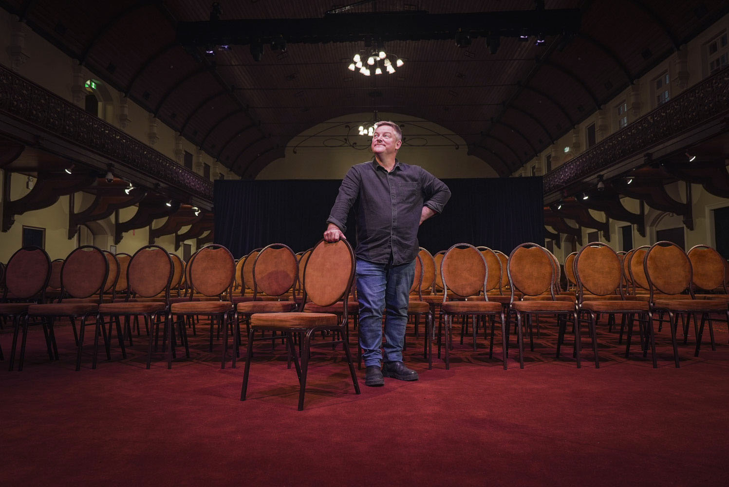 A man with grey hair sits on a chair in front of a theatre auditorium