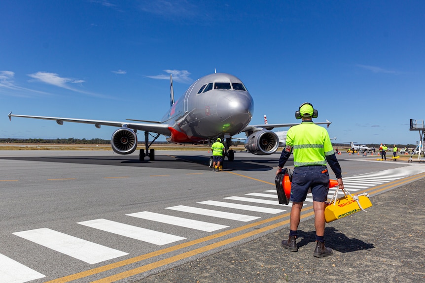 A plane taxiing on a runway, while a worker stands wearing high-vis top and holds equipment.