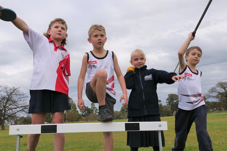 Four kids in a field, with one holding a discuss, another a shot put, the third a javelin and the fourth stepping over a hurdle