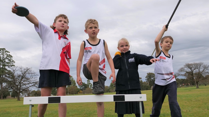 Four kids in a field, with one holding a discuss, another a shot put, the third a javelin and the fourth stepping over a hurdle