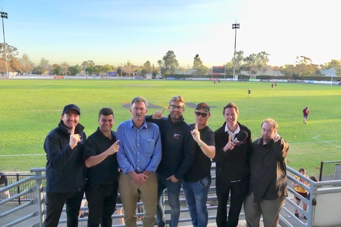 A group of men stand in a football stand with a football ground behind him. 