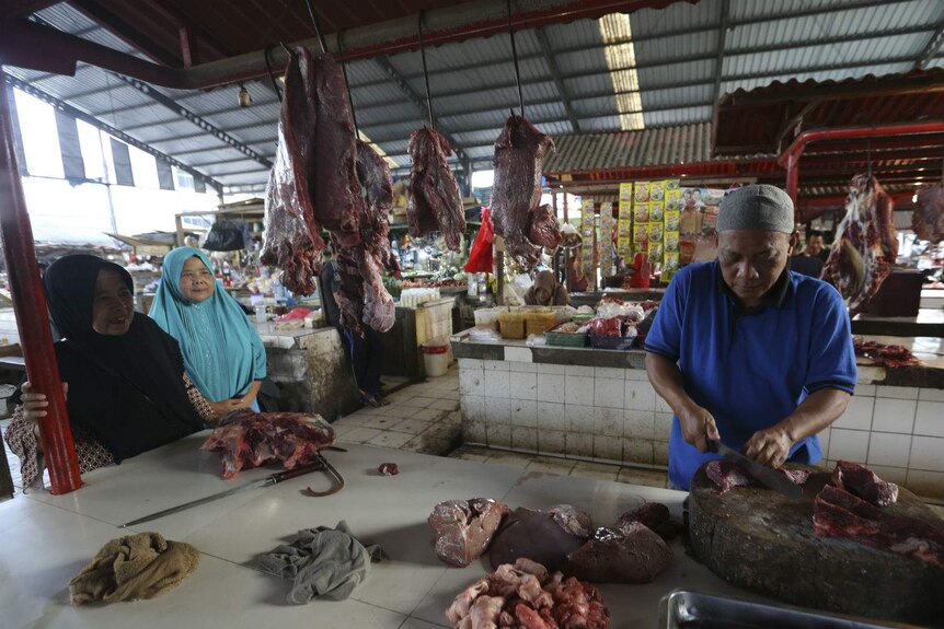 Two women in headscarves chat to a man in an Islamic cap as he carves meat at a market butcher's stall.