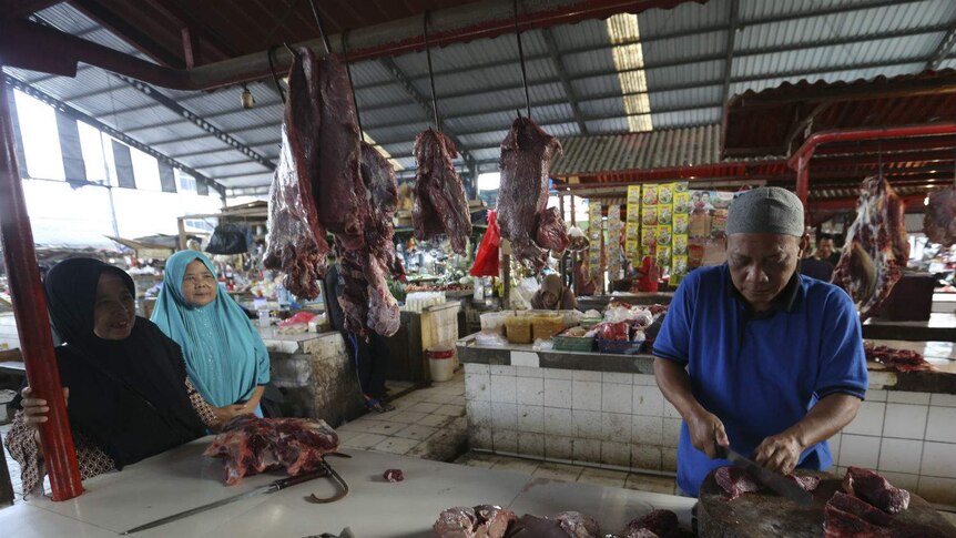 Two women in headscarves chat to a man in an Islamic cap as he carves meat at a market butcher's stall.