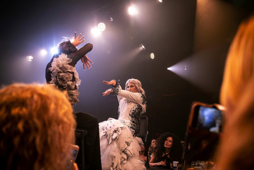 Two performers sit opposite each other on stage, moving their hands in the air, while audience members look on.