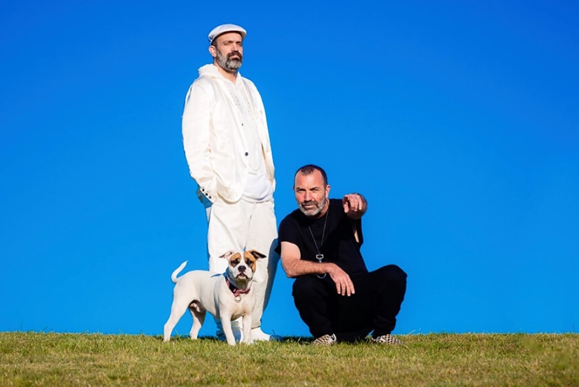 Johnny Seymour stands wearing white, Paul Mac crouches wearing black, they are on a green grassy hill with a white dog