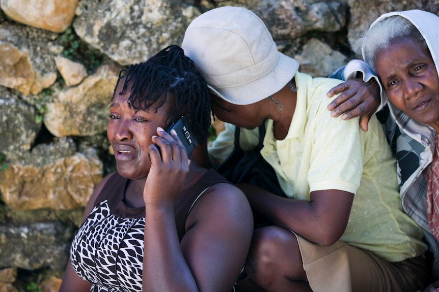 A woman on her phone sheds tears as a young man with a hat on stands behind her and an elderly woman rests her arm on him.