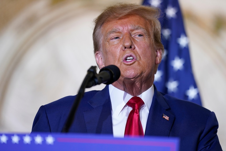 Donald Trump in blue suit and red tie speaking at podium with US flag in the background.