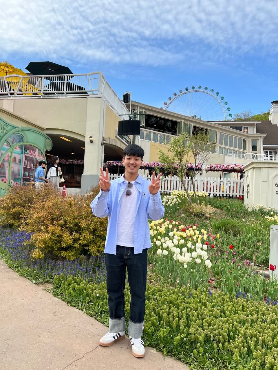 A young man wearing blue shirt stands in front of a garden. 